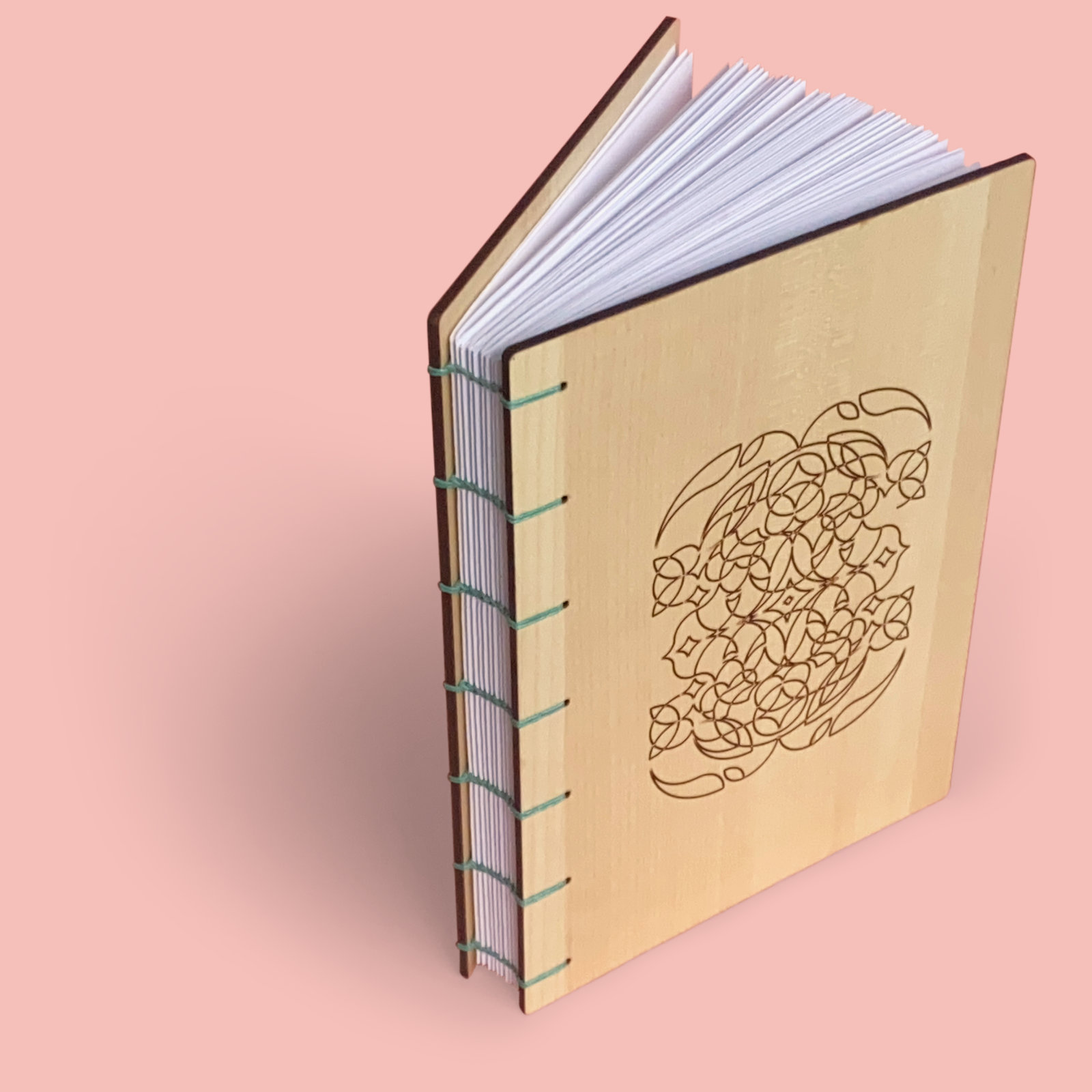 A book with maple wood covers stands partly open against a pale pink background. There is a unique linear design engraved on the front. The spine of the book is exposed and you can see each signature. Teal 4ply waxed Irish linen thread has been used to bind the pages using the coptic binding technique.