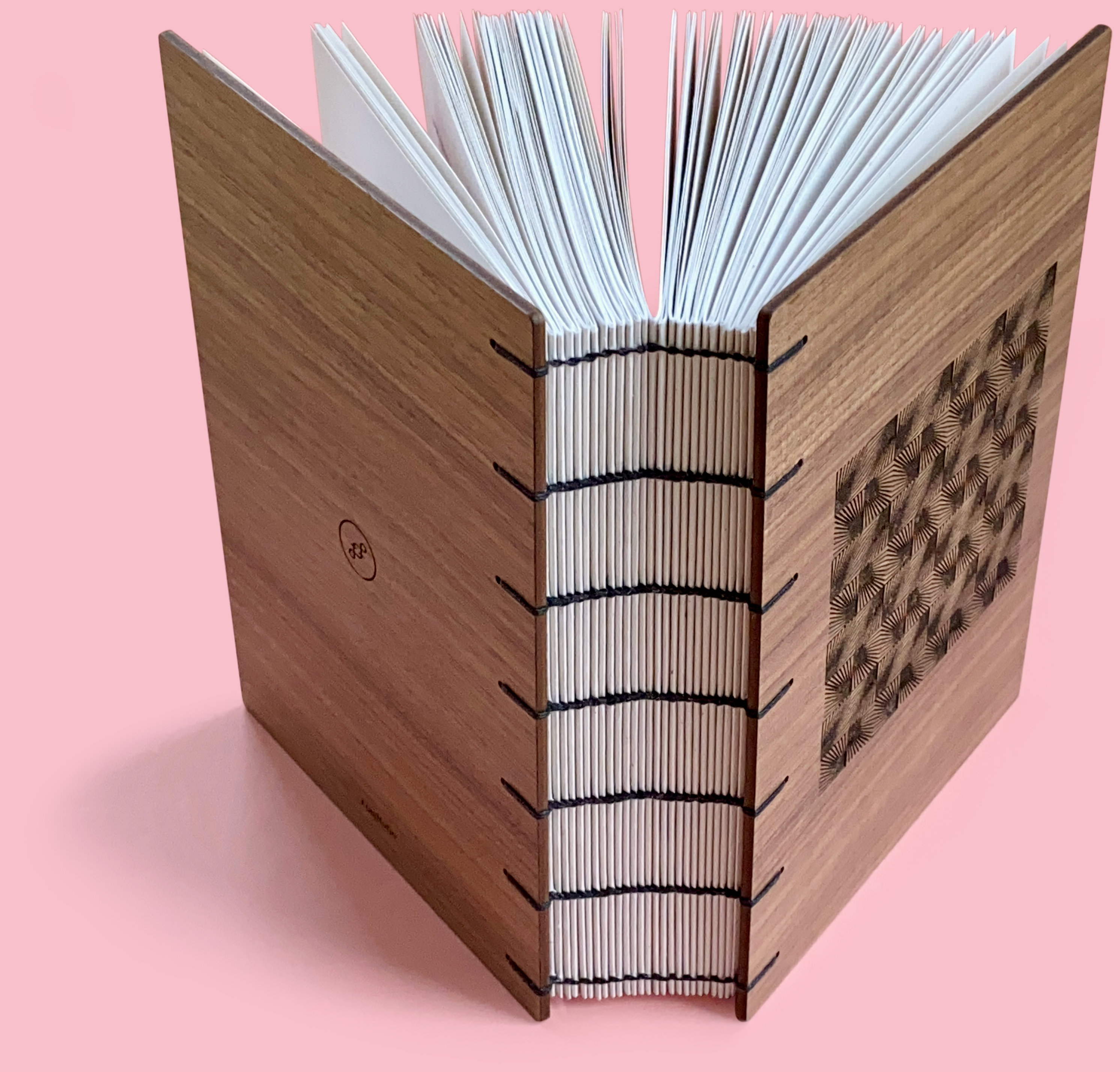 A book with walnut wood covers stands open against a pastel pink background. There is a unique geometric pattern engraved on the front. The spine of the book is exposed and you can see each signature. Black 4ply waxed Irish linen thread has been used to bind the pages using the coptic binding technique.