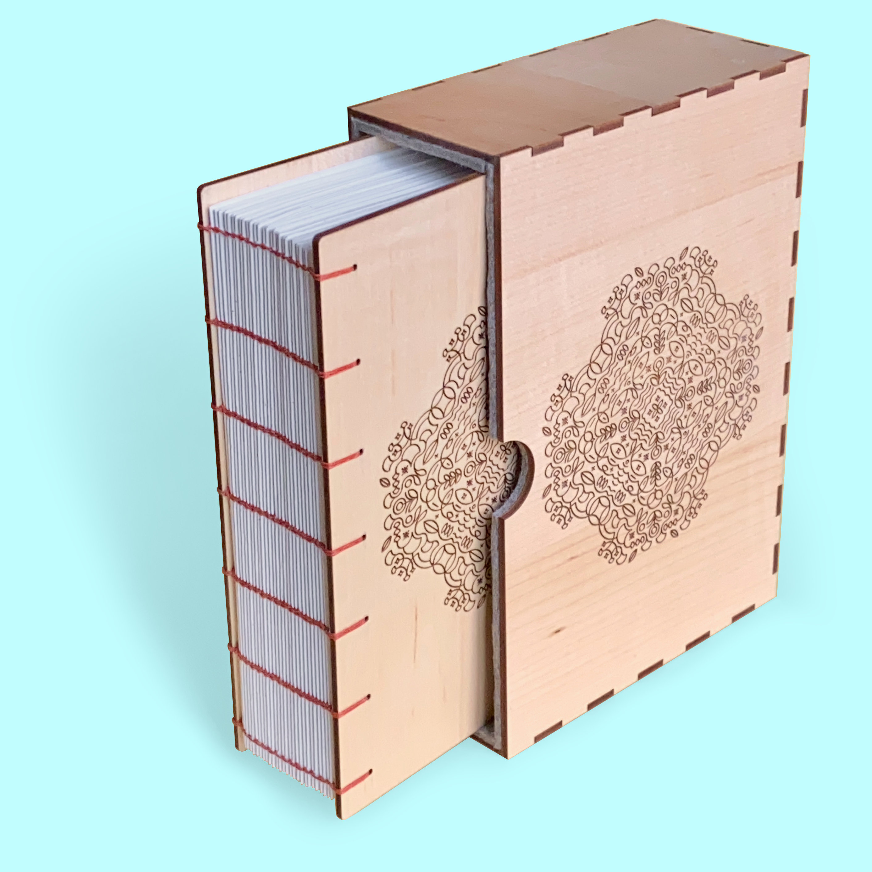 Against a pale blue background a book with maple wood covers is emerging from a custom maple slipcase. There is a unique mandala style pattern engraved on the front. The spine of the book is exposed and you can see each signature. Rusty red 4ply waxed Irish linen thread has been used to bind the pages using the coptic binding technique. The slipcase uses finger joint construction which creates a pleasant pattern along the edges.