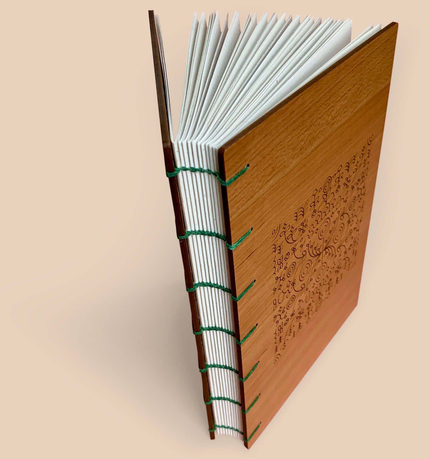 A book with cherry wood covers stands partly open against a pale orange background. There is a unique pattern engraved on the front. The spine of the book is exposed and you can see each signature. Emerald green 4ply waxed Irish linen thread has been used to bind the pages using the coptic binding technique.